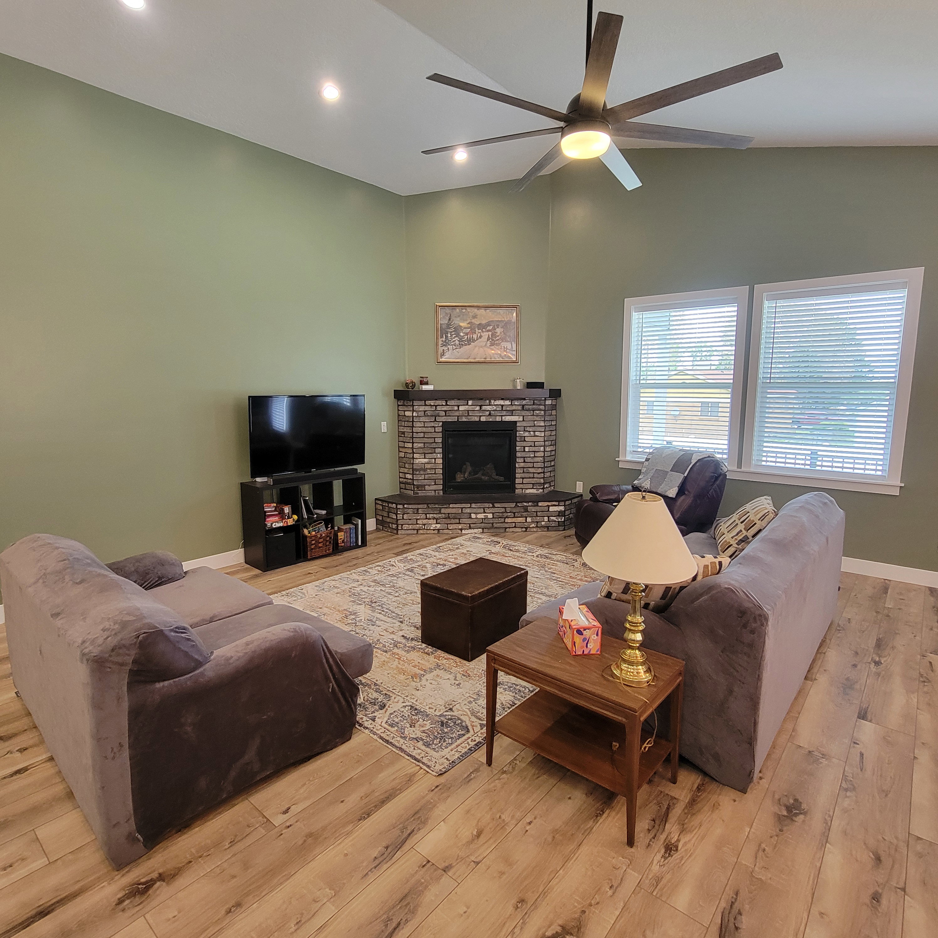 Spacious living area featuring a corner fireplace with brick surround and wood mantel, light wood-finish flooring, and a vaulted ceiling with recessed lighting and a ceiling fan
