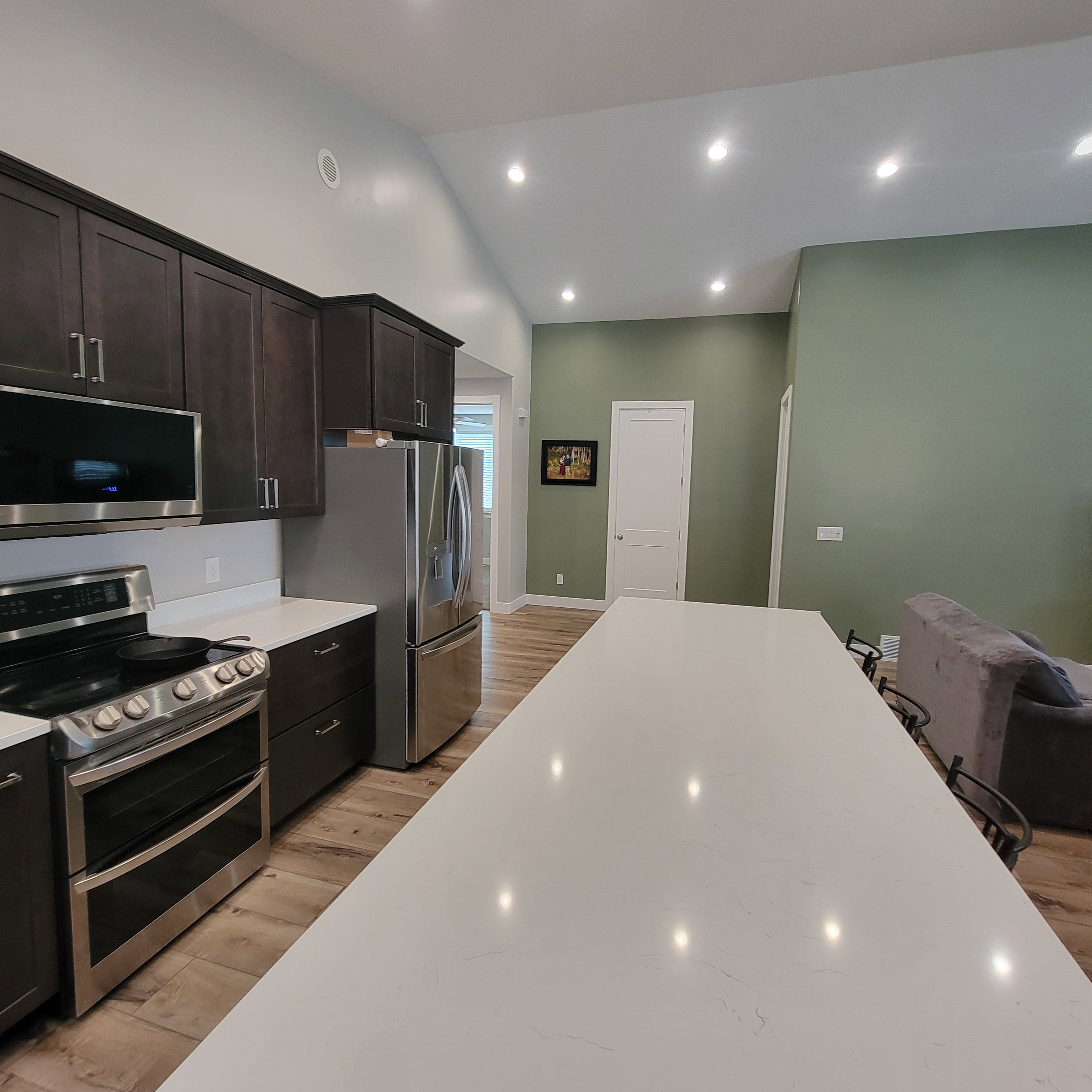 Kitchen featuring dark wood cabinetry, stainless steel appliances, a long white countertop island, wood-finish flooring, and vaulted ceilings with recessed lighting