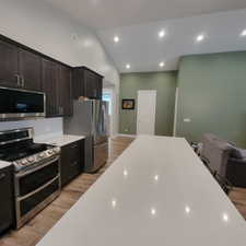 Kitchen featuring dark wood cabinetry, stainless steel appliances, a long white countertop island, wood-finish flooring, and vaulted ceilings with recessed lighting