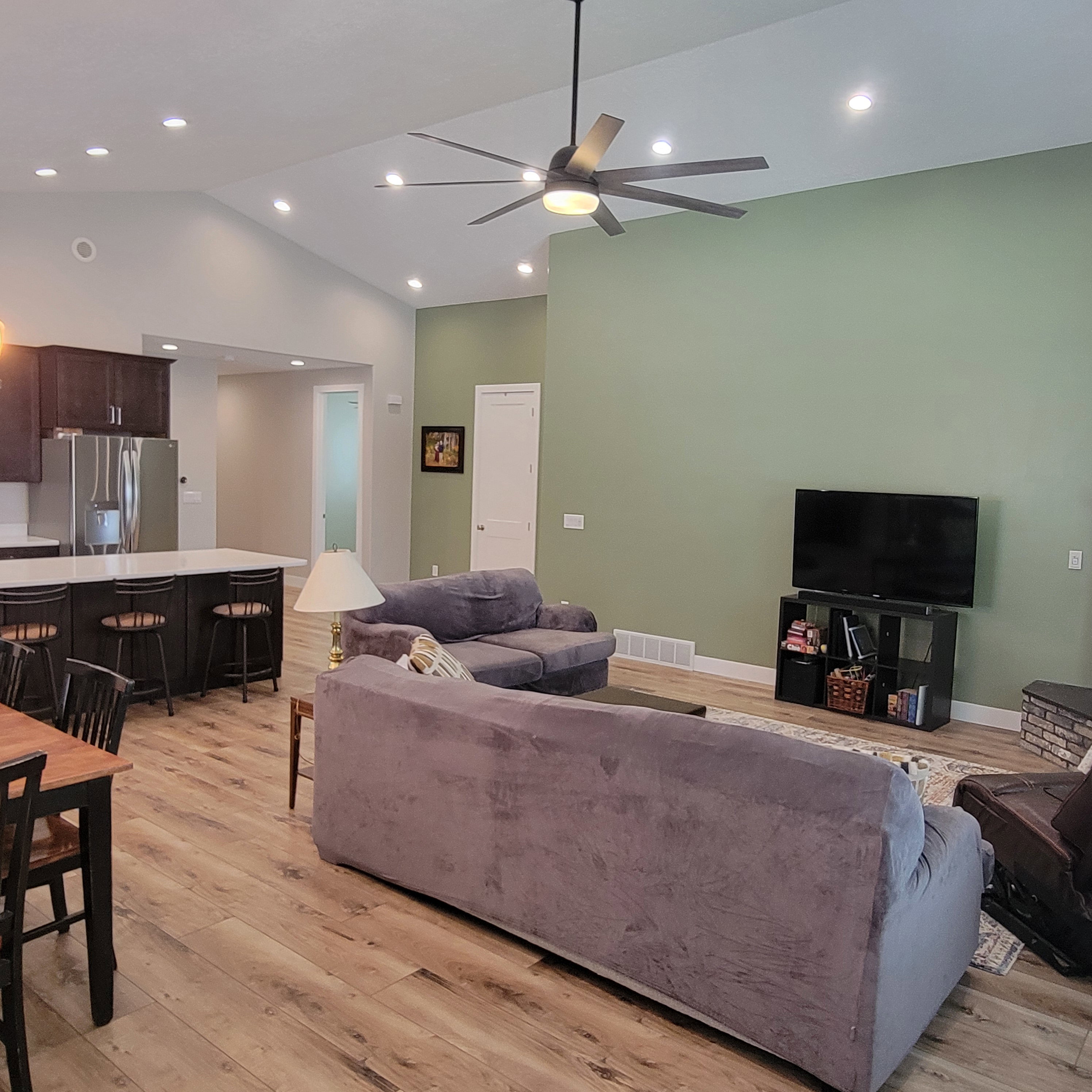 Open-concept living area featuring vaulted ceilings with recessed lighting, a large ceiling fan, wood-finish flooring, and a kitchen island with seating