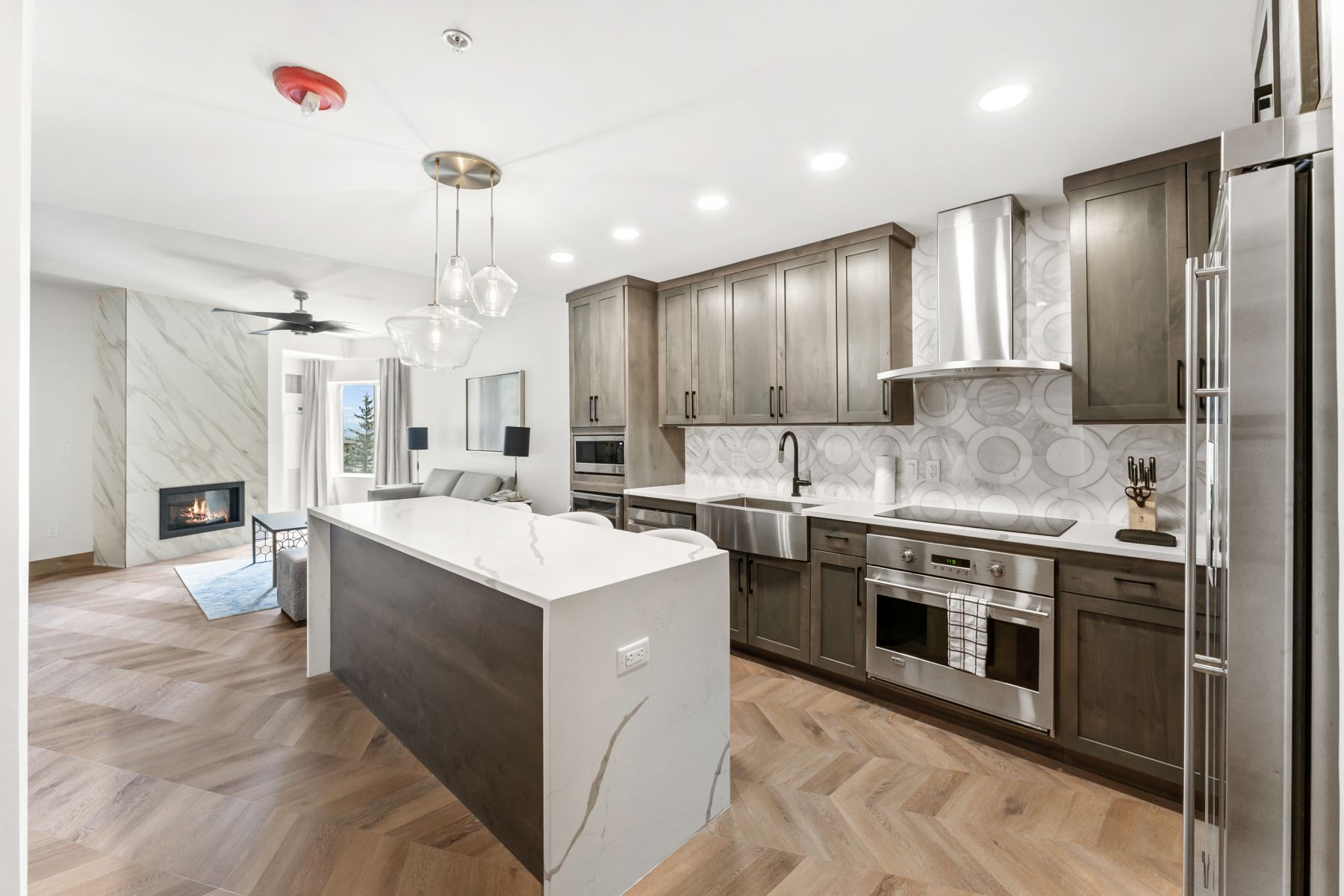 Open-concept living space featuring a herringbone wood-finish floor, a central kitchen island with a white countertop, dark wood-finish cabinetry, stainless steel appliances, and a marble-clad fireplace