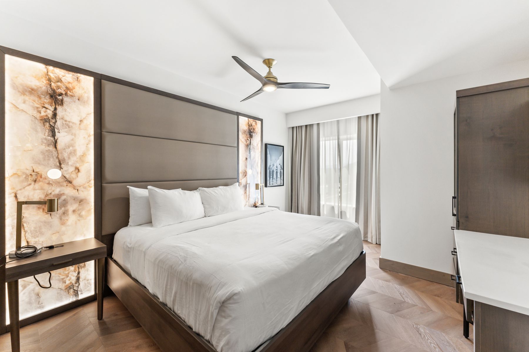 Bedroom featuring a padded headboard with illuminated onyx accent panels, wood-finish herringbone flooring, and a ceiling fan