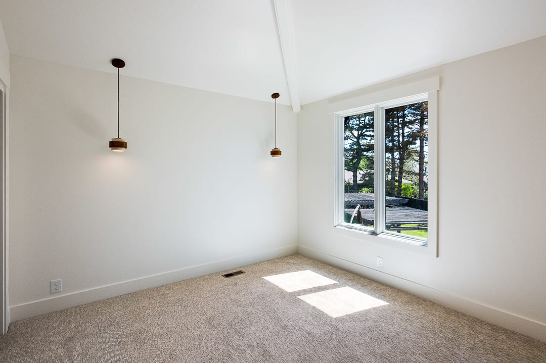 Vaulted ceiling room featuring a large window, neutral wall paint, plush carpeting, and two contemporary pendant lights with wood-finish accents
