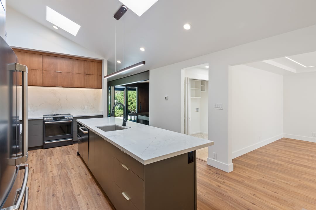 Modern kitchen featuring a vaulted ceiling with skylights, wood-finish flooring, a central island with an integrated sink, stainless steel appliances, and a sophisticated two-tone cabinetry design