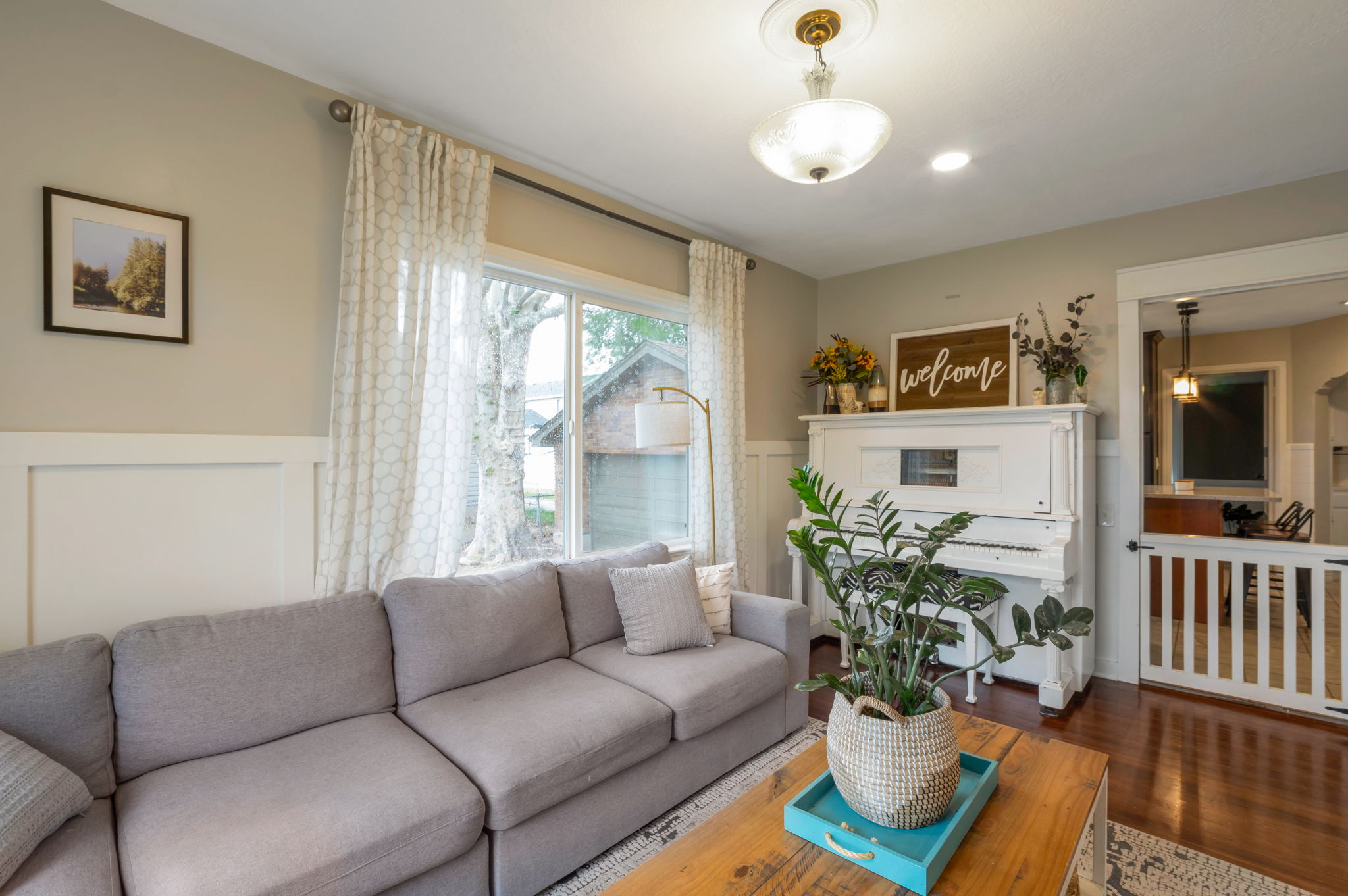 Bright living space featuring warm wood-finish flooring, wainscoting, and large windows with privacy drapes