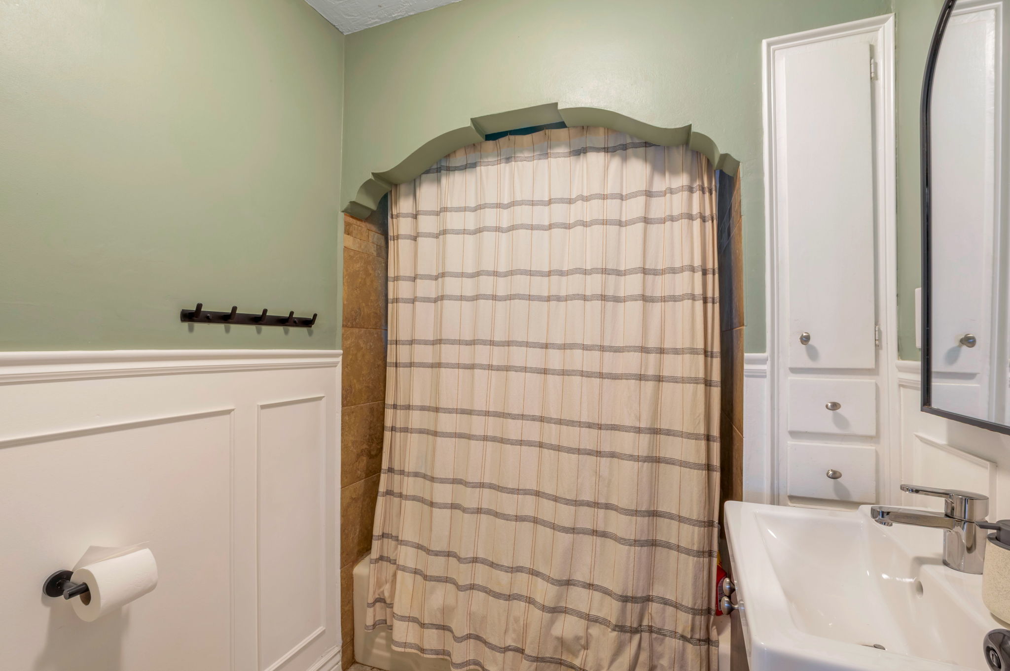 Bathroom featuring wainscoting, an arched shower surround with tile, a pedestal sink with a modern faucet, and built-in cabinetry