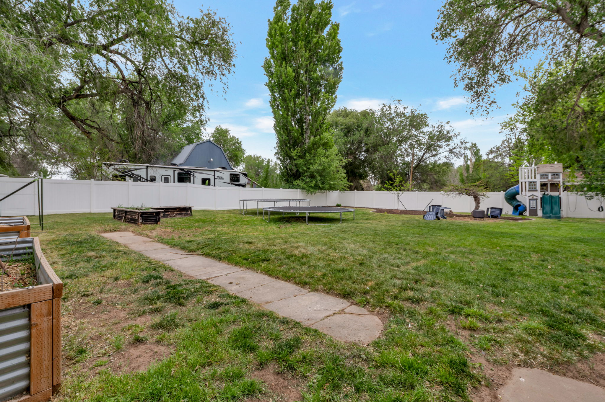 Expansive backyard featuring a concrete pathway, two raised garden beds with wood and corrugated metal accents, a white privacy fence, and mature trees