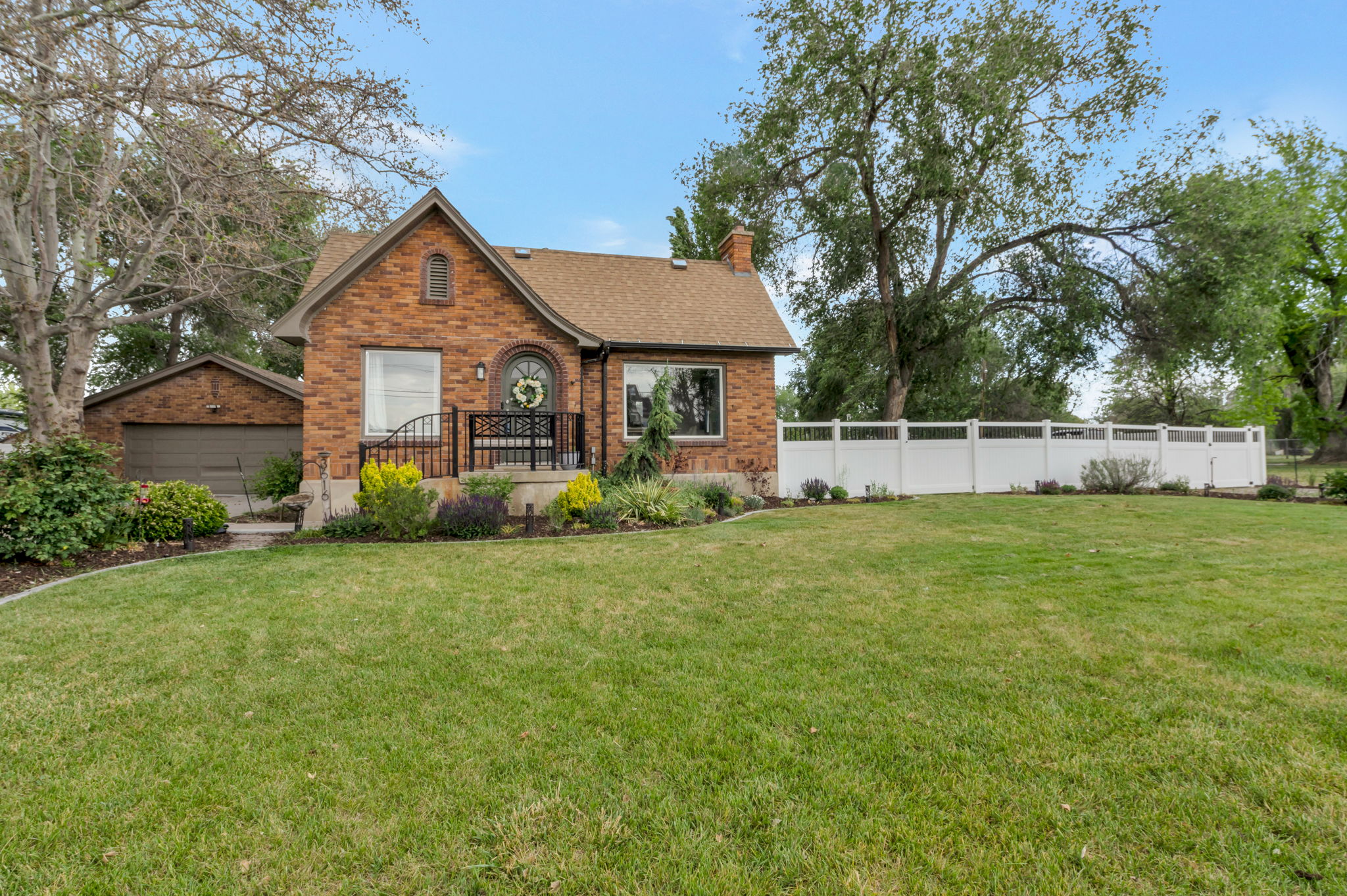 Traditional brick residence featuring an attached garage, a front porch with wrought iron railing, a white picket fence, a meticulously maintained lawn, and mature trees