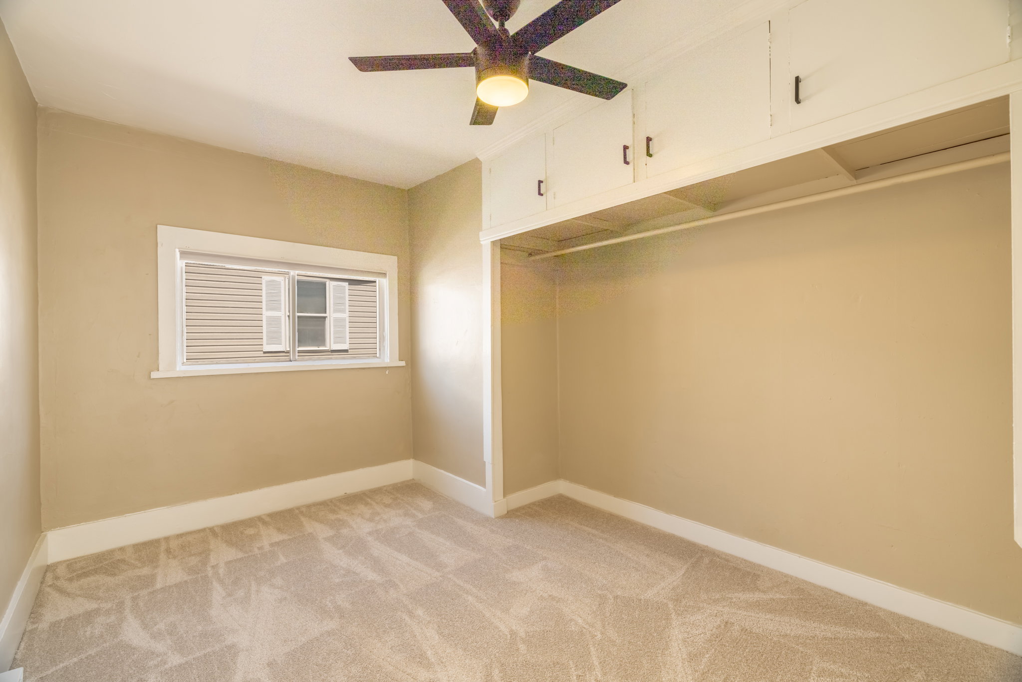 Room featuring neutral-toned walls, light-colored patterned carpeting, a white-framed window with shutters, a ceiling fan with integrated lighting, and built-in overhead cabinetry with an open closet rod