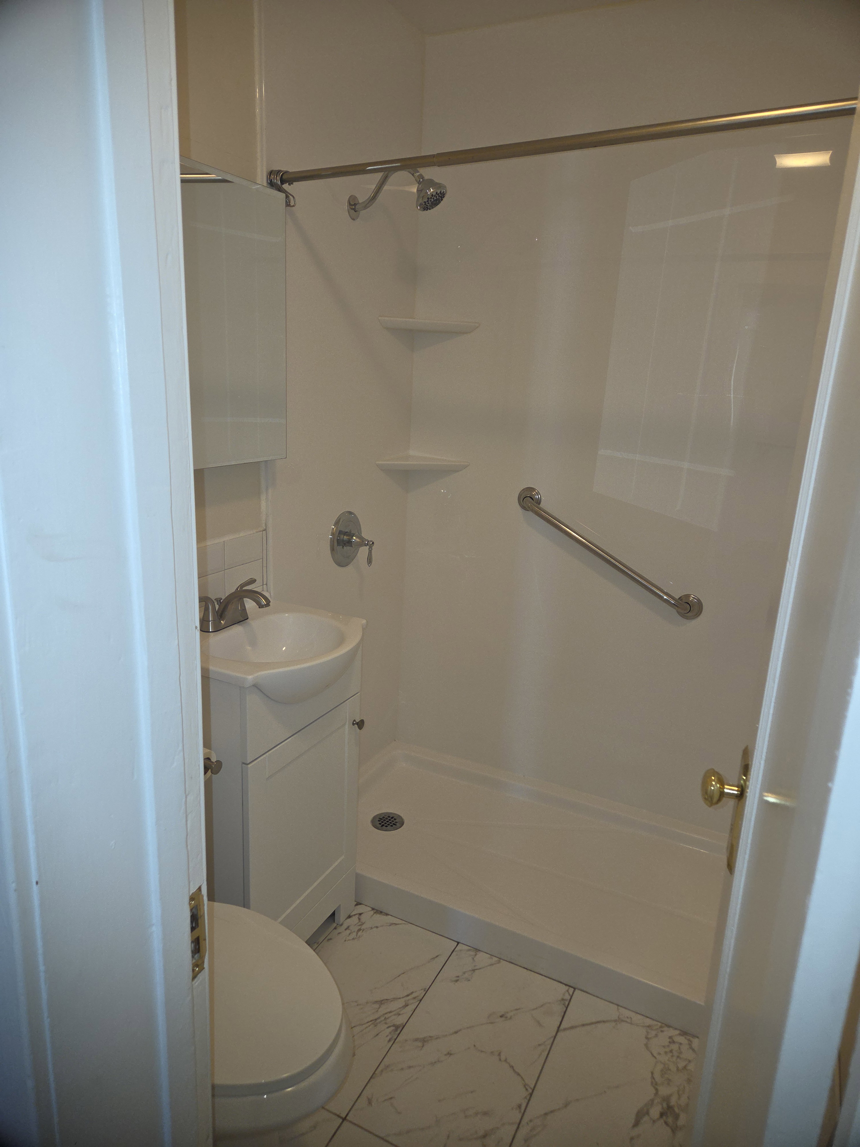 Compact bathroom featuring a white shower stall with built-in shelving and a grab bar, a white pedestal sink with brushed nickel faucet, a white toilet, and marble-look tile flooring