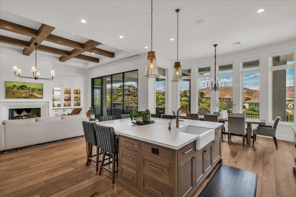 Spacious open-concept living area featuring a large kitchen island with a farmhouse sink, wood-finish flooring, a fireplace, exposed ceiling beams, and expansive windows