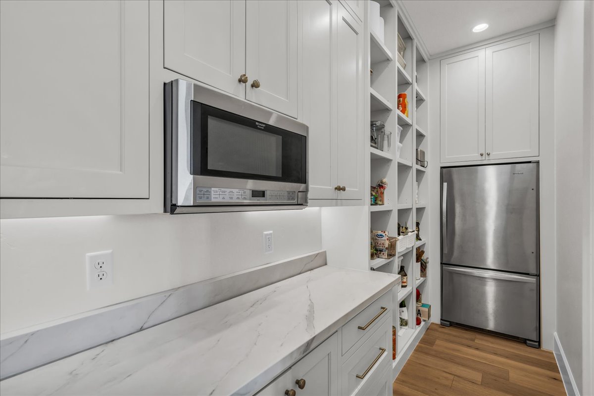 Built-in stainless steel microwave over a light-toned countertop, white cabinetry with brass-toned hardware, under-cabinet lighting, and expansive built-in shelving