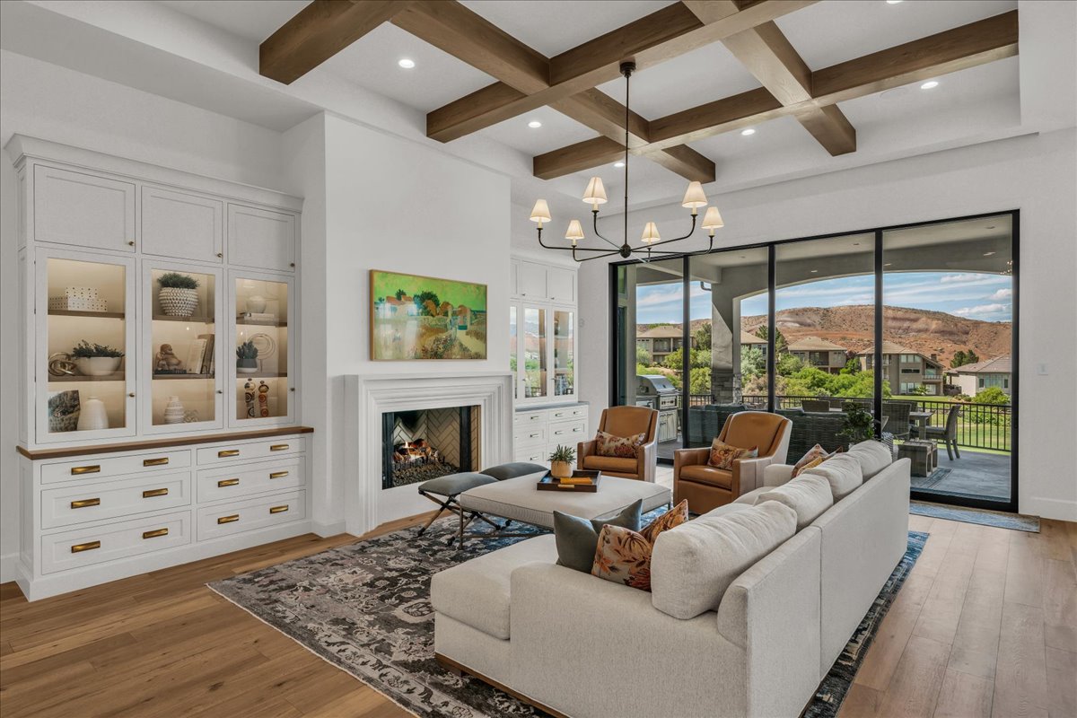 Living area featuring wood-finish flooring, a coffered ceiling with exposed wood beams, a fireplace with a white mantel, and built-in white cabinetry with glass display doors