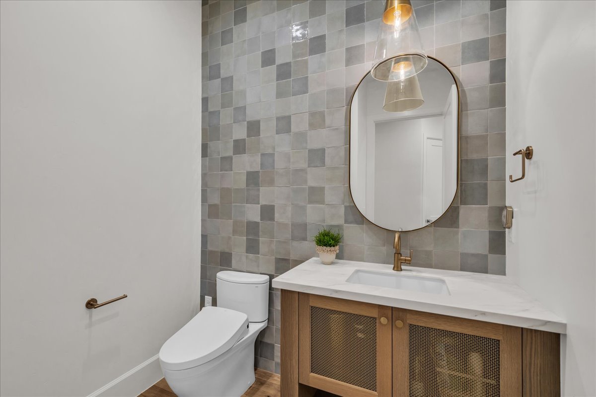 Powder room featuring a mosaic tile accent wall, wood-finish vanity with mesh cabinet doors, integrated sink, and an oval mirror with brass-tone fixtures