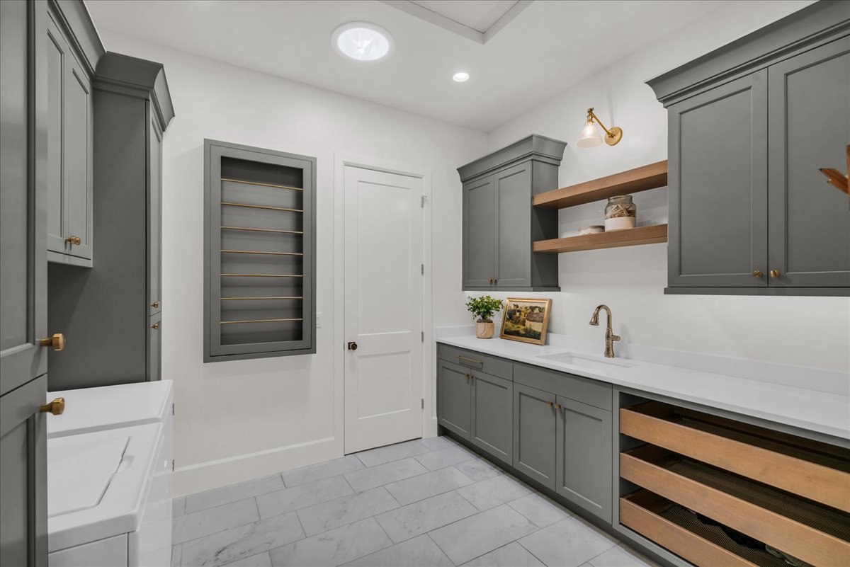 Spacious laundry room featuring extensive built-in cabinetry, durable tile flooring, a large sink basin with a gooseneck faucet, integrated drying racks, and recessed lighting