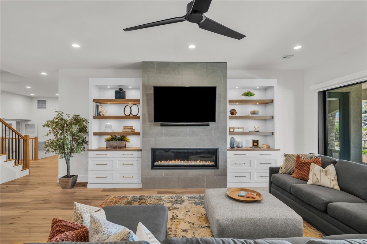 Living area featuring a floor-to-ceiling tiled fireplace, built-in shelving with integrated lighting, wood-finish flooring, and a modern ceiling fan