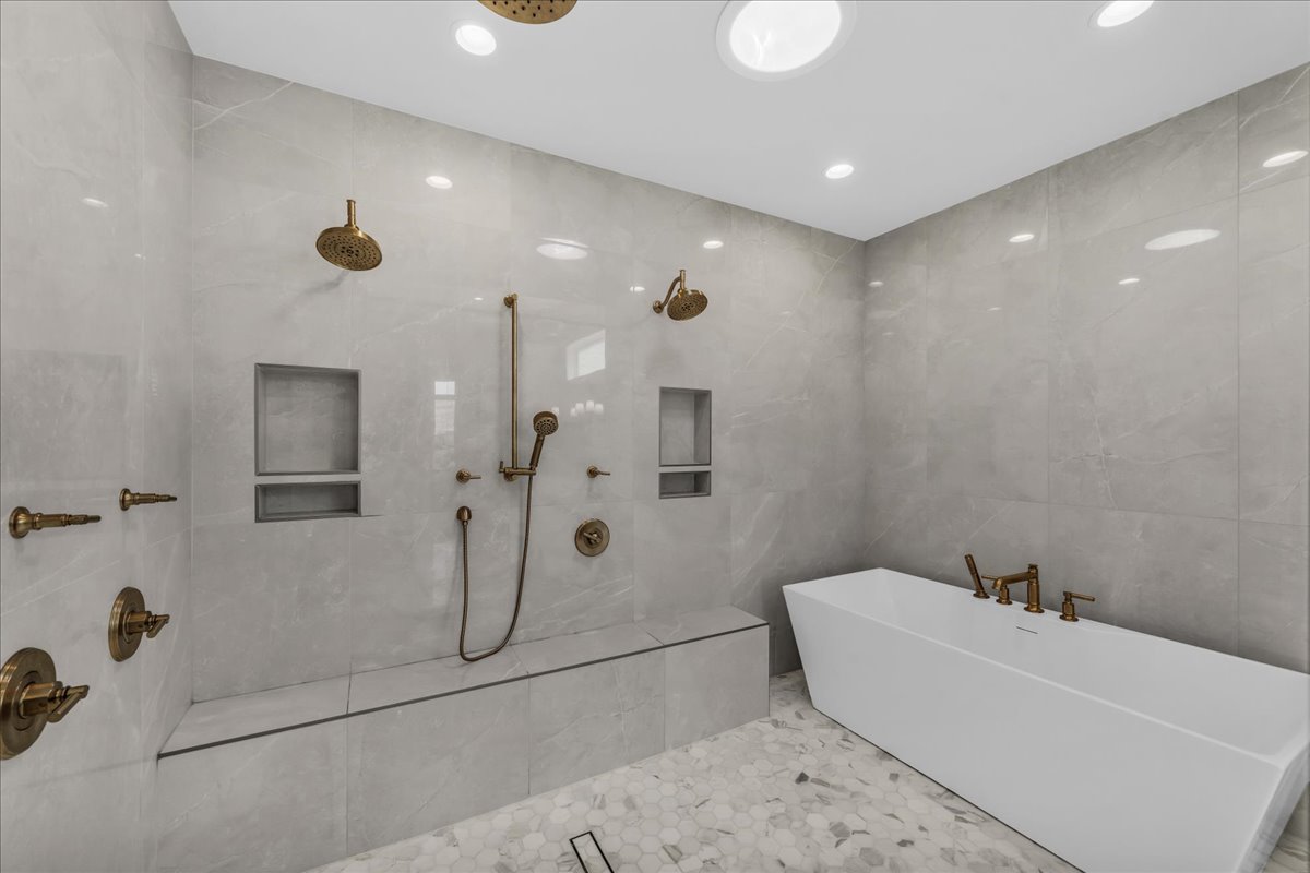 Spacious wet room featuring a freestanding soaking tub, dual rain shower heads, handheld shower fixture, built-in shower bench, and integrated shower niches