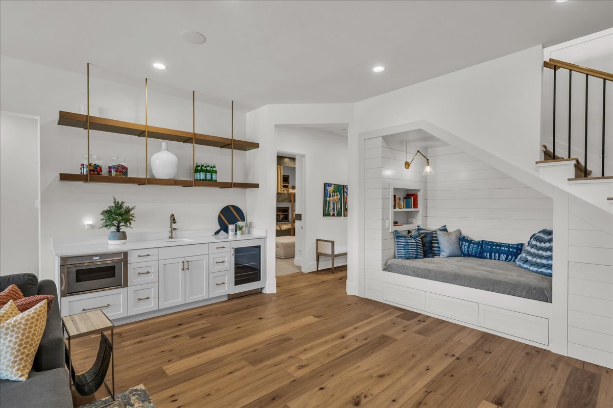 Hardwood flooring throughout a versatile space featuring a built-in wet bar with under-counter microwave and beverage cooler, and a custom window seat with integrated shelving and wall sconce lighting
