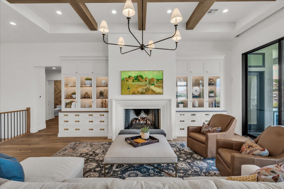 Spacious living area featuring exposed wood ceiling beams, a decorative fireplace with a mantel, built-in cabinetry with glass-front uppers and solid lower drawers, and large format windows