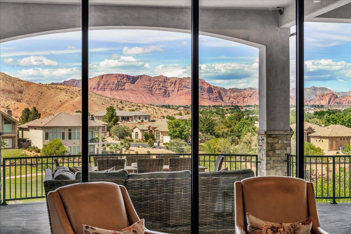 Expansive sliding glass doors open to an outdoor living area with a stone pillar and black metal railing, featuring distant red rock formations and verdant landscape