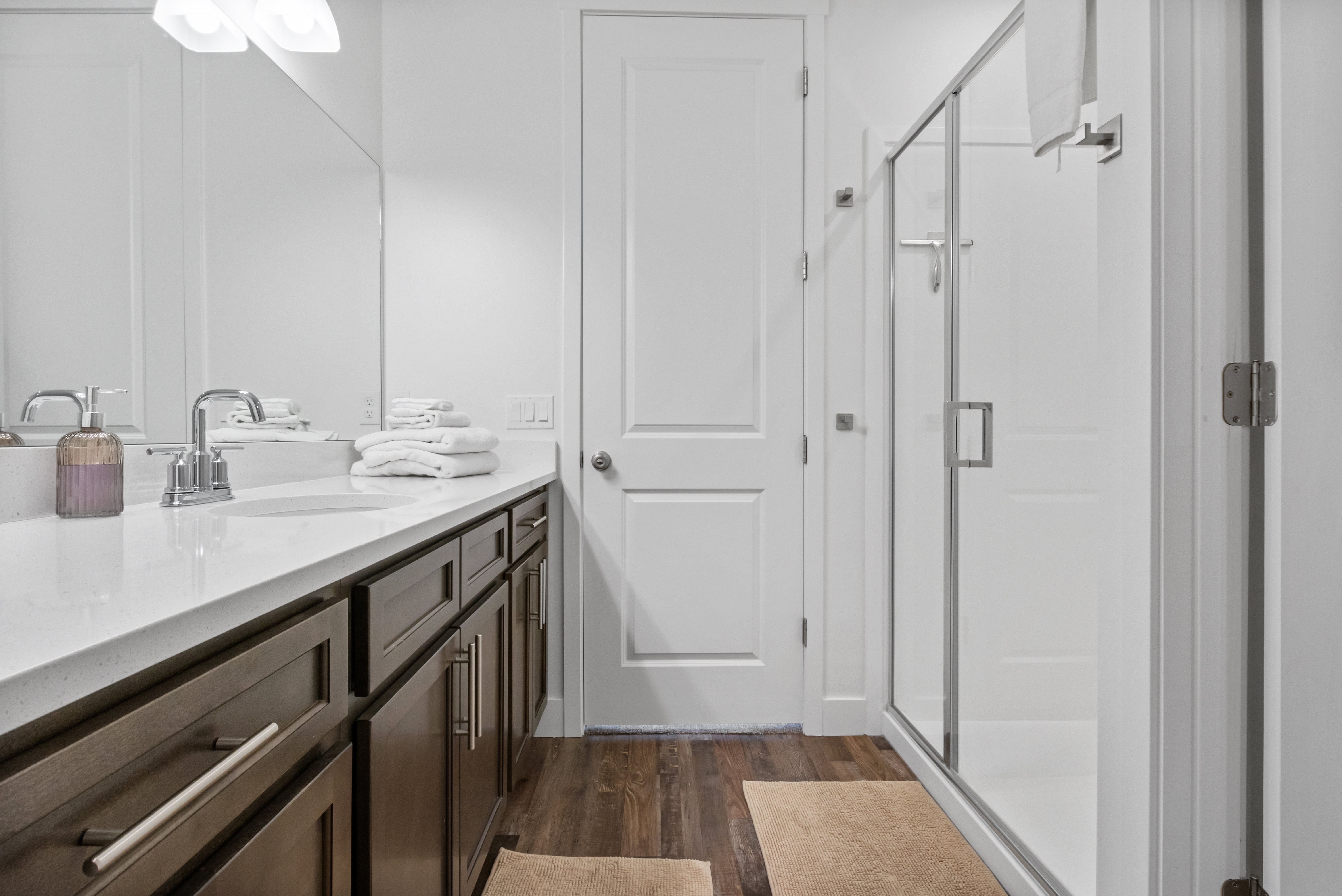 Bathroom featuring a wood-finish floor, dark wood cabinetry with brushed nickel hardware, a white countertop with an undermount sink, and a glass-enclosed shower with a brushed nickel frame