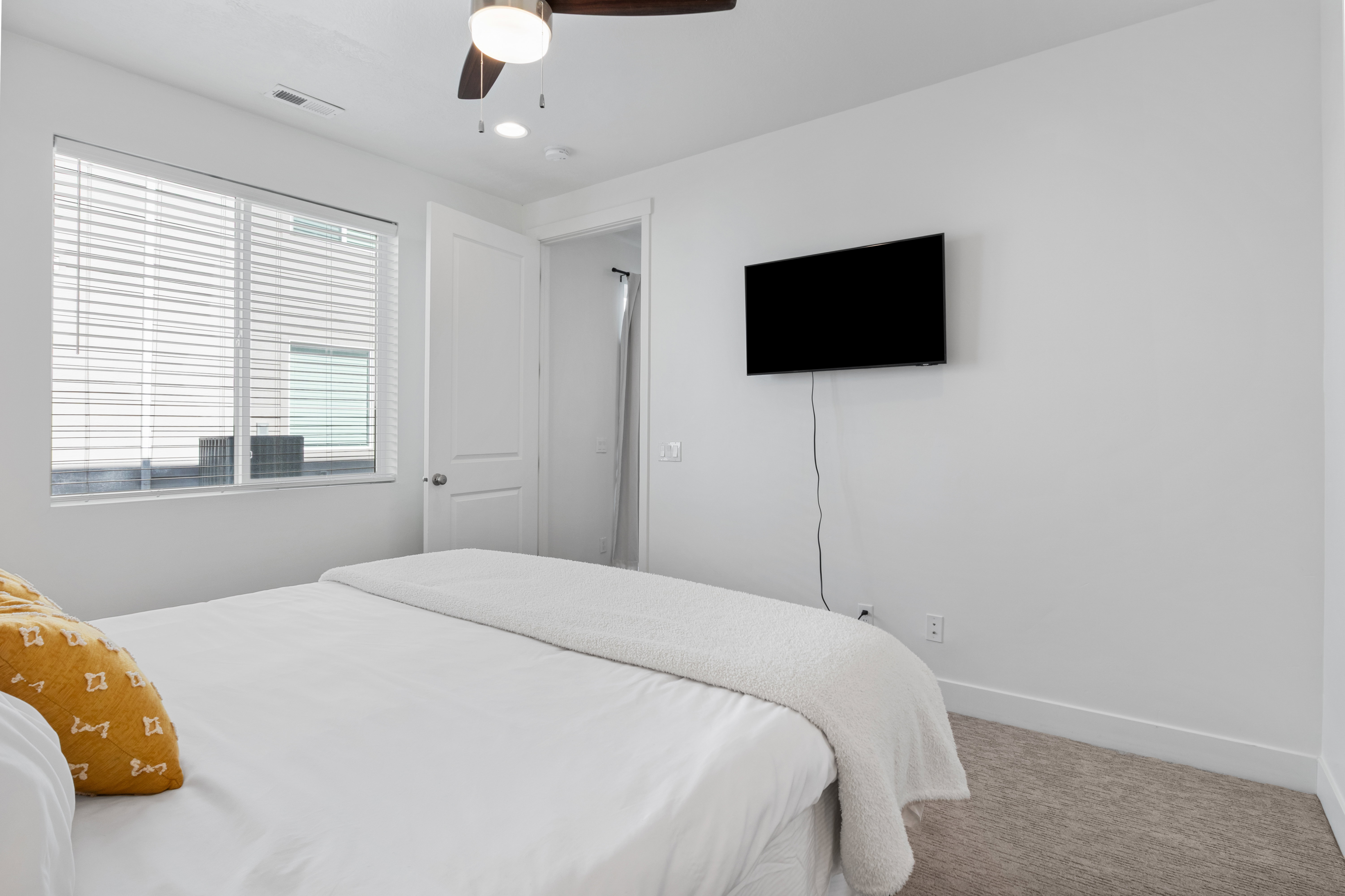 Bright bedroom with large window featuring white horizontal blinds, a ceiling fan with integrated lighting, and neutral tone carpeting