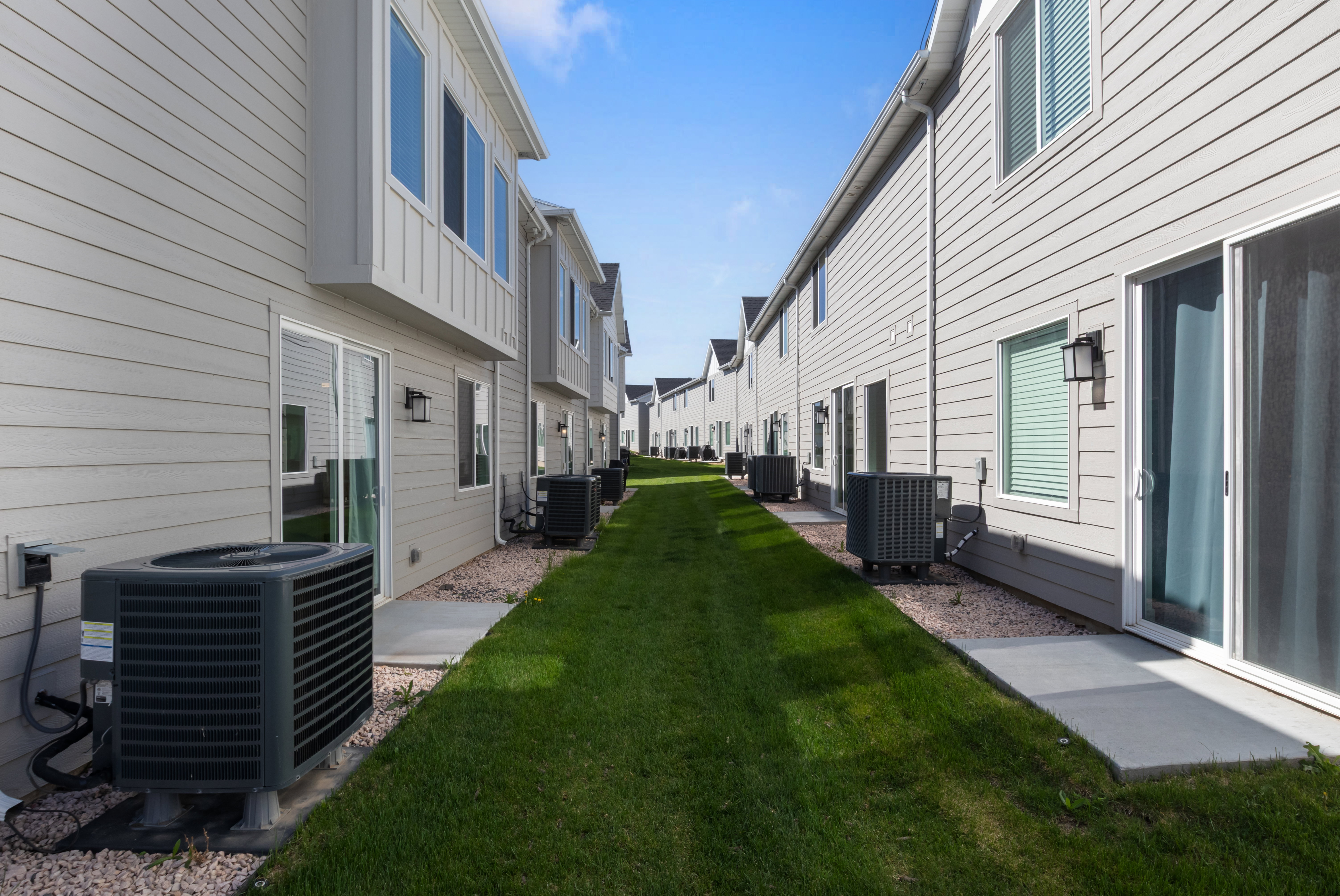 Row of modern townhomes featuring light-colored siding, individual patios, sliding glass doors, and manicured grass pathways