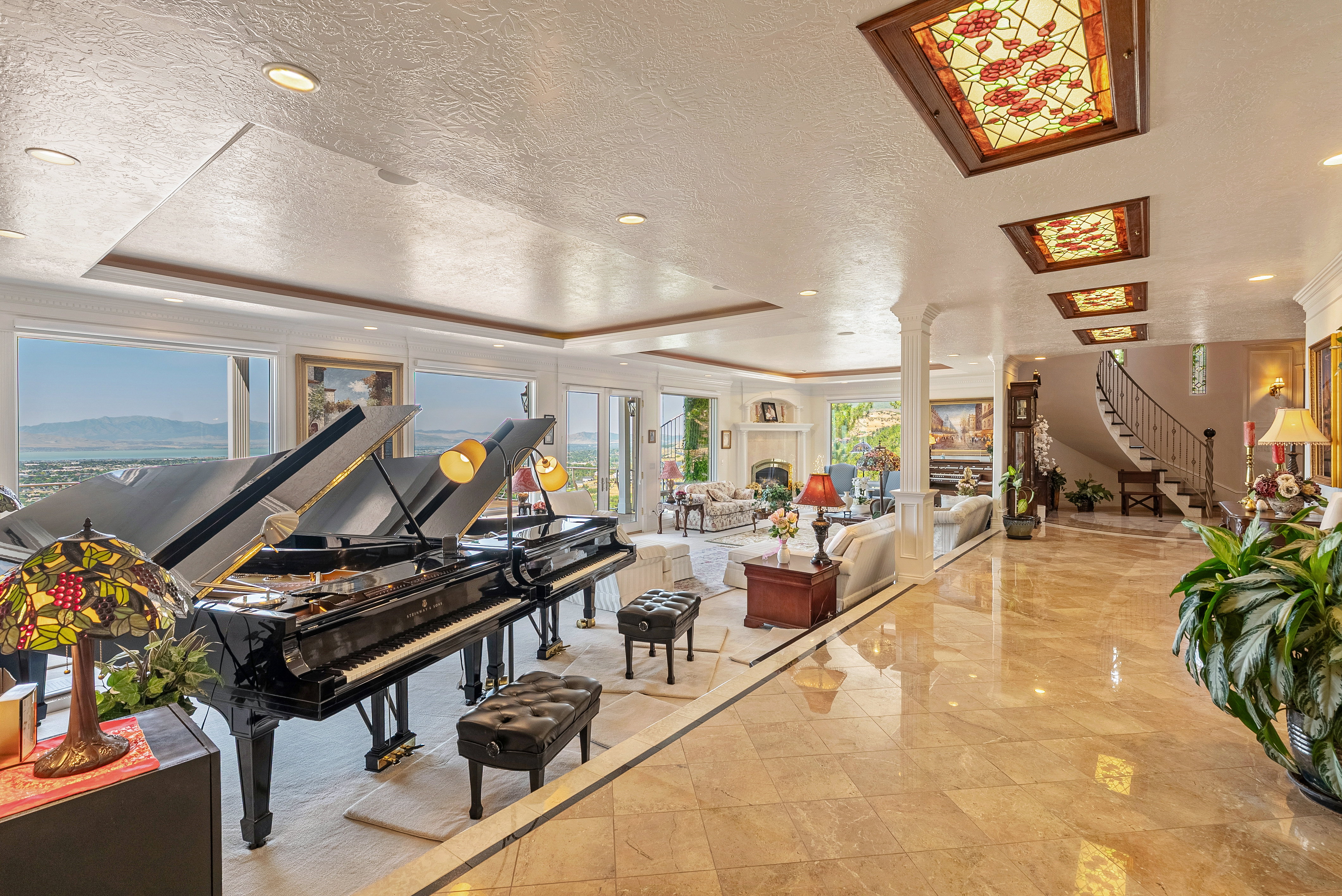 Expansive interior featuring polished tile flooring, a grand staircase with wrought iron railing, decorative stained-glass ceiling panels, recessed lighting, and a wall of windows showcasing mountain scenery