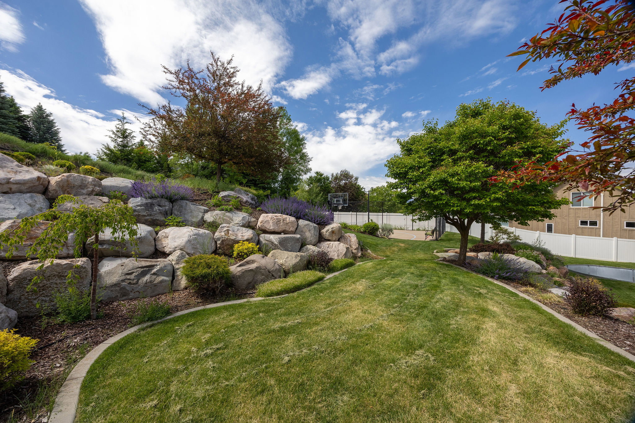 Expansive terraced backyard featuring a manicured lawn, established trees, a rock retaining wall, diverse landscaping, and a fenced perimeter