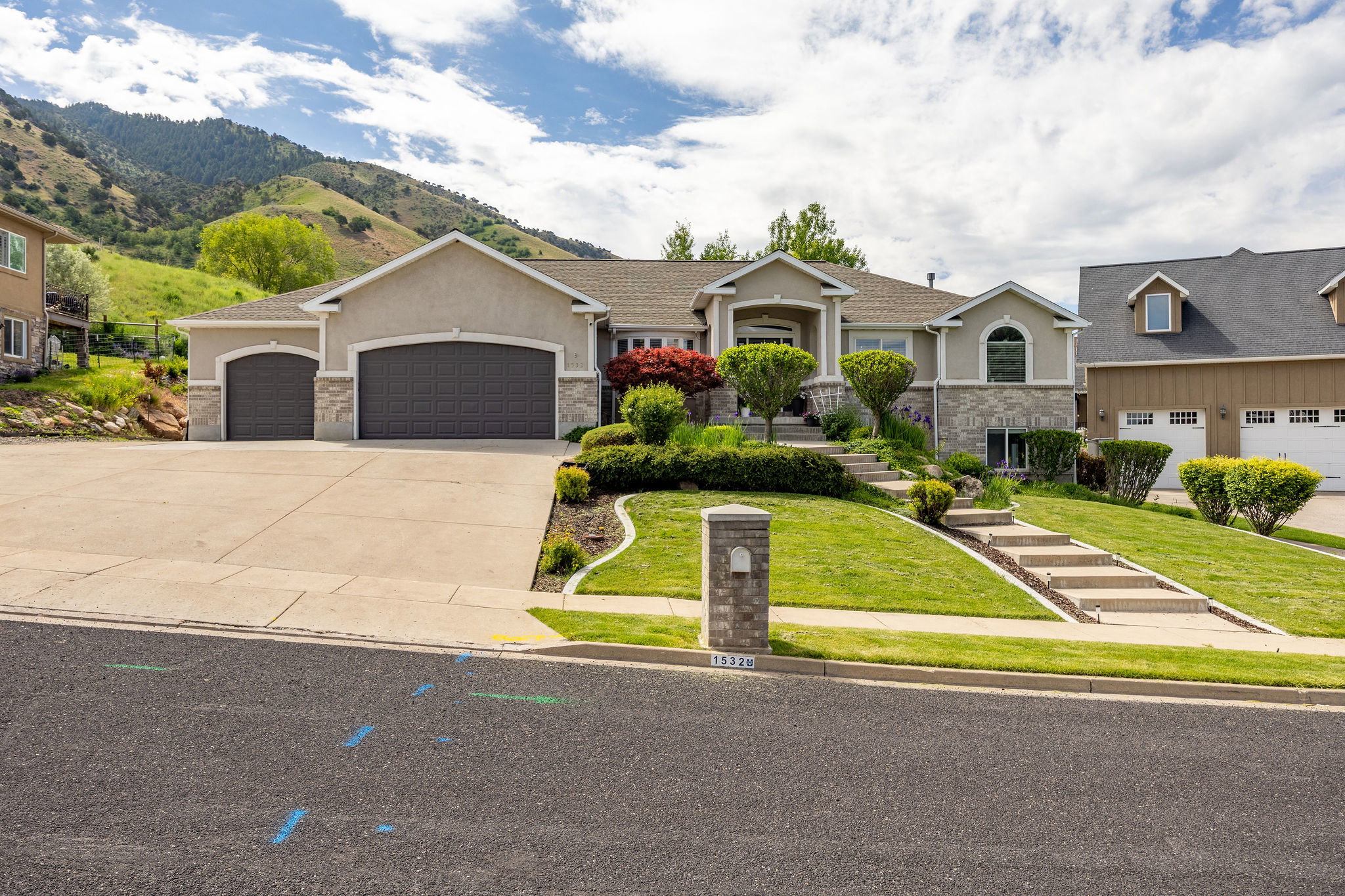 Stucco and stone facade property featuring a three-car garage, tiered landscaping with mature shrubs, concrete driveway, and a paved walkway leading to the entrance
