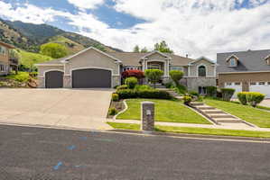 Stucco and stone facade property featuring a three-car garage, tiered landscaping with mature shrubs, concrete driveway, and a paved walkway leading to the entrance