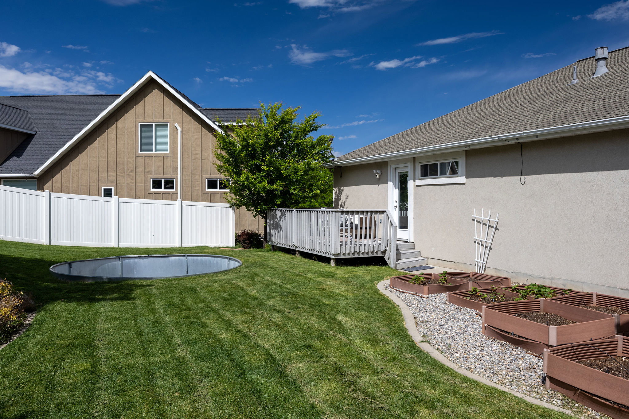 Expansive backyard featuring a green lawn, white vinyl fencing, a built-in metal ring, a multi-level deck, and raised garden beds