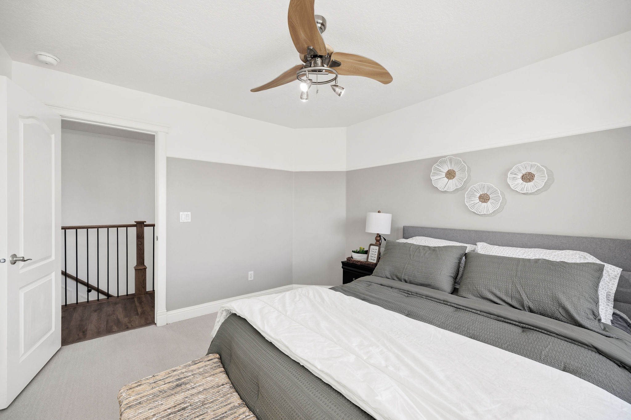 Bedroom featuring a ceiling fan with wood-finish blades, light gray painted walls, and an open doorway leading to a staircase with a black metal railing and wood-finish flooring