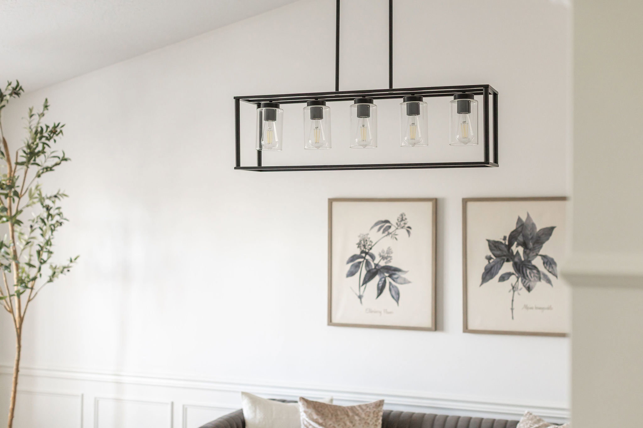 Linear chandelier with a black frame and clear glass shades, illuminating a space with white walls and classic wainscoting