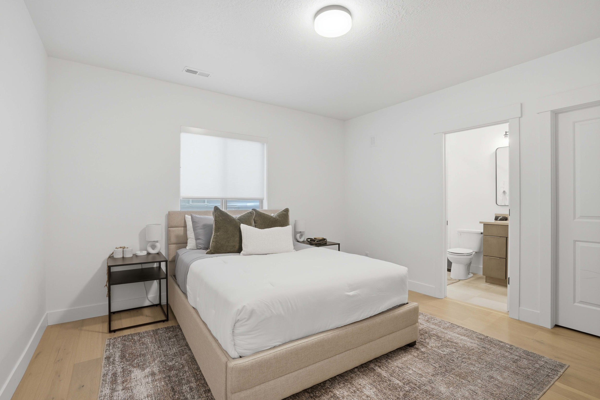 Bedroom featuring light wood-finish flooring, a contemporary window with a roller shade, and an en-suite bathroom entry