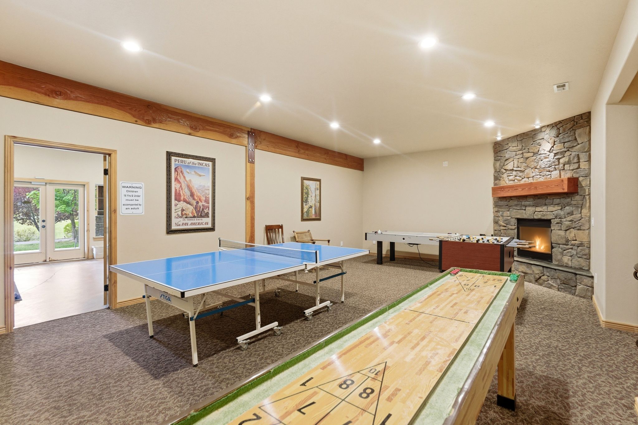 Spacious recreation room featuring recessed lighting, exposed wood beams, and a stone-clad fireplace with a wood mantel