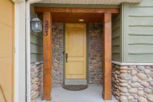 Entryway featuring a natural wood door, stacked stone facade, exposed wood beams, and a concrete porch