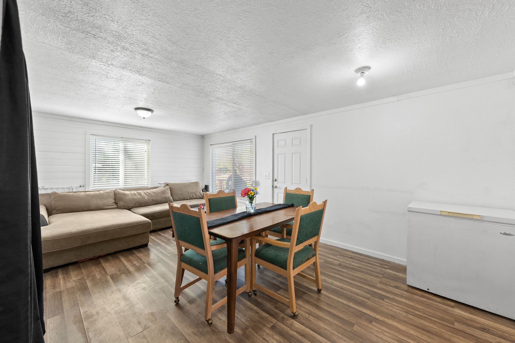 Open-concept living space featuring wood-finish flooring, white shiplap-style accent wall, and multiple windows with blinds
