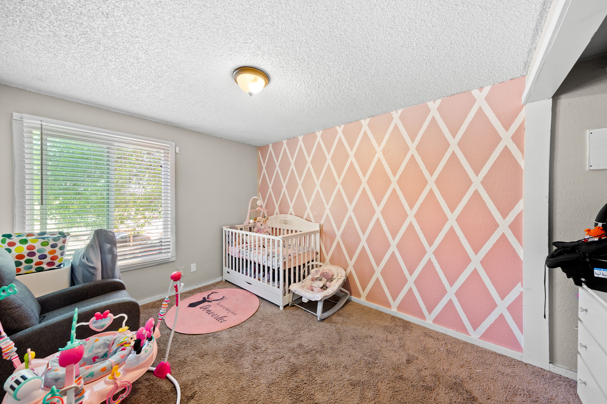 Spacious interior room featuring a decorative accent wall with a geometric diamond pattern, neutral-toned carpeting, and a large window with horizontal blinds