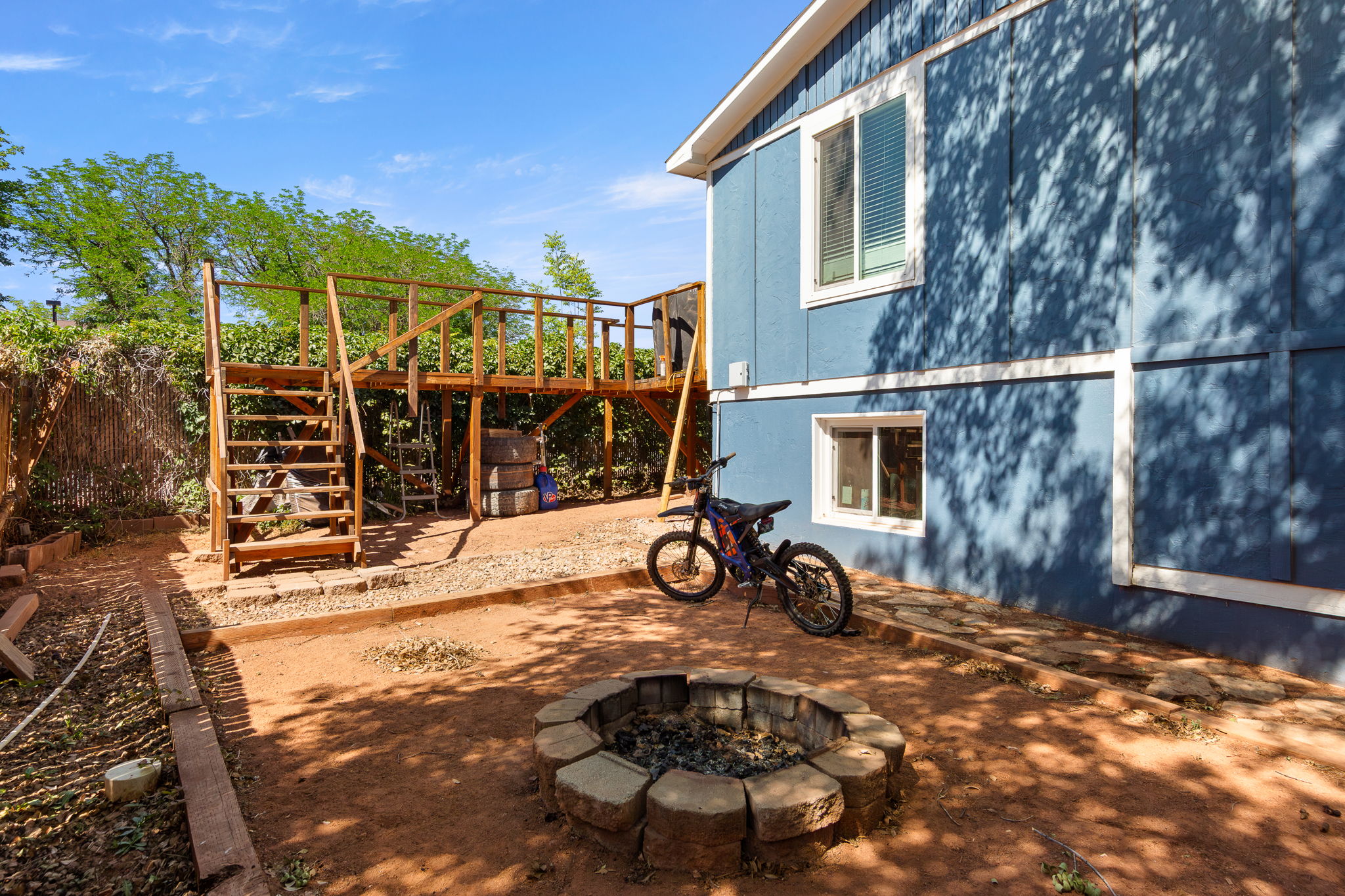 Backyard featuring a built-in fire pit with stone block surround, a multi-level wooden deck structure with stairs, and a fence with natural screening