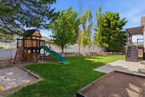 Open backyard featuring a lush lawn, mature trees, concrete patio, built-in planter box, built-in sandbox, and playground