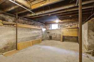 Unfinished basement featuring concrete flooring, exposed wooden joists, and a visible window for natural light