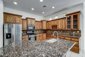 Kitchen featuring a granite-look island with an undermount sink, stainless steel appliances, wood cabinetry, tile backsplash, and recessed lighting