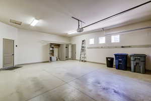 Spacious garage featuring concrete flooring, multiple fluorescent lighting fixtures, a garage door opener mechanism, built-in shelving, and three small windows providing natural light