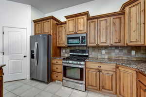 Kitchen featuring stainless steel appliances, granite-style countertops, wood cabinetry with crown molding, a tiled backsplash, and light-toned floor tiles