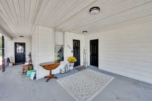 Covered front porch featuring two black paneled entry doors, white siding, a window with white trim, and overhead lighting