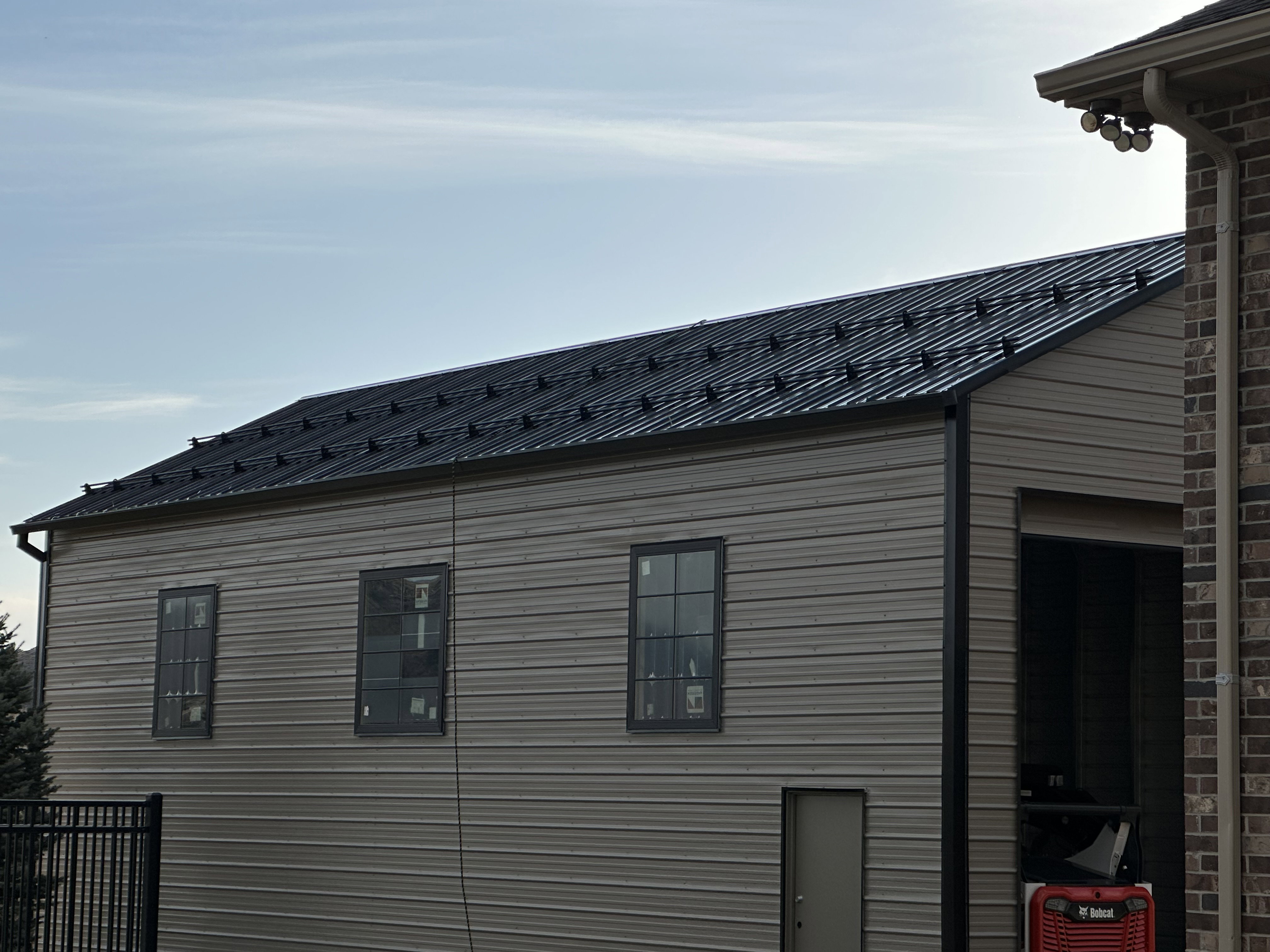 Detached outbuilding with a dark metal standing seam roof, light beige horizontal siding, and multiple dark-framed windows
