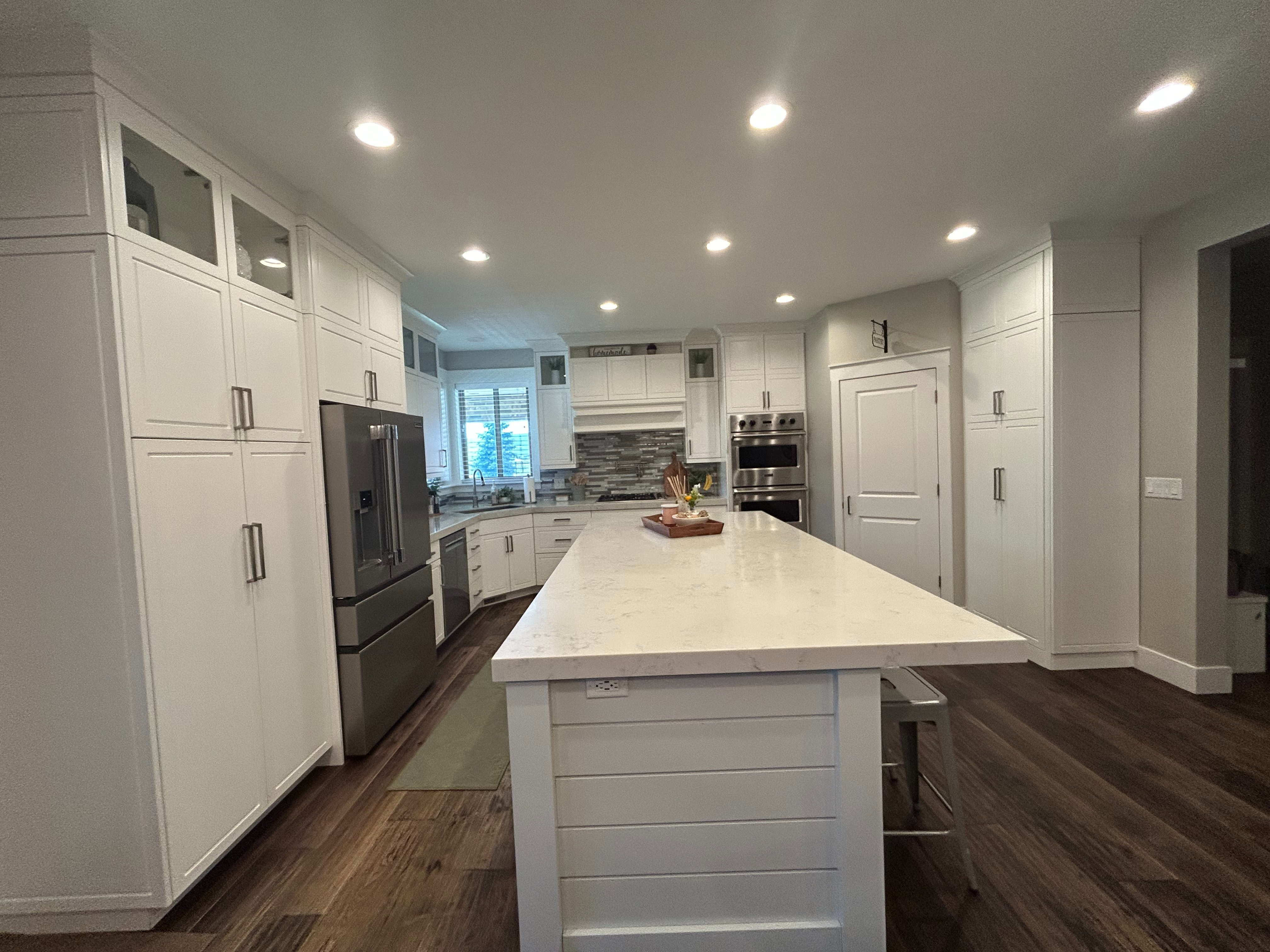 Expansive kitchen featuring a large central island with a light-toned countertop, extensive white cabinetry, wood-finish flooring, recessed lighting, and stainless steel appliances