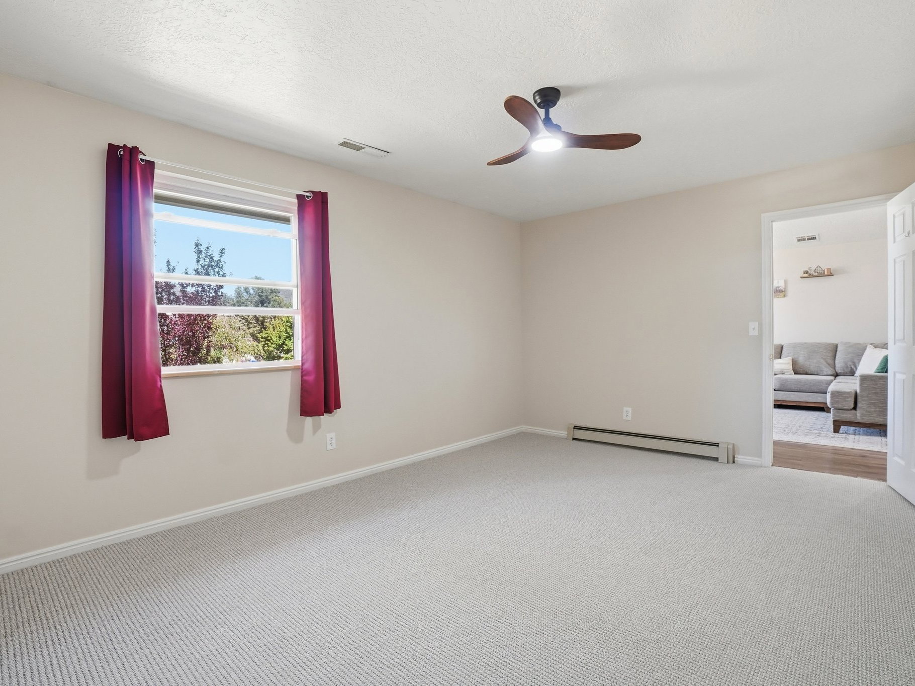 Spacious room featuring light neutral wall paint, textured carpet flooring, a window offering exterior foliage, a ceiling fan with integrated lighting, and an open doorway revealing an adjacent room