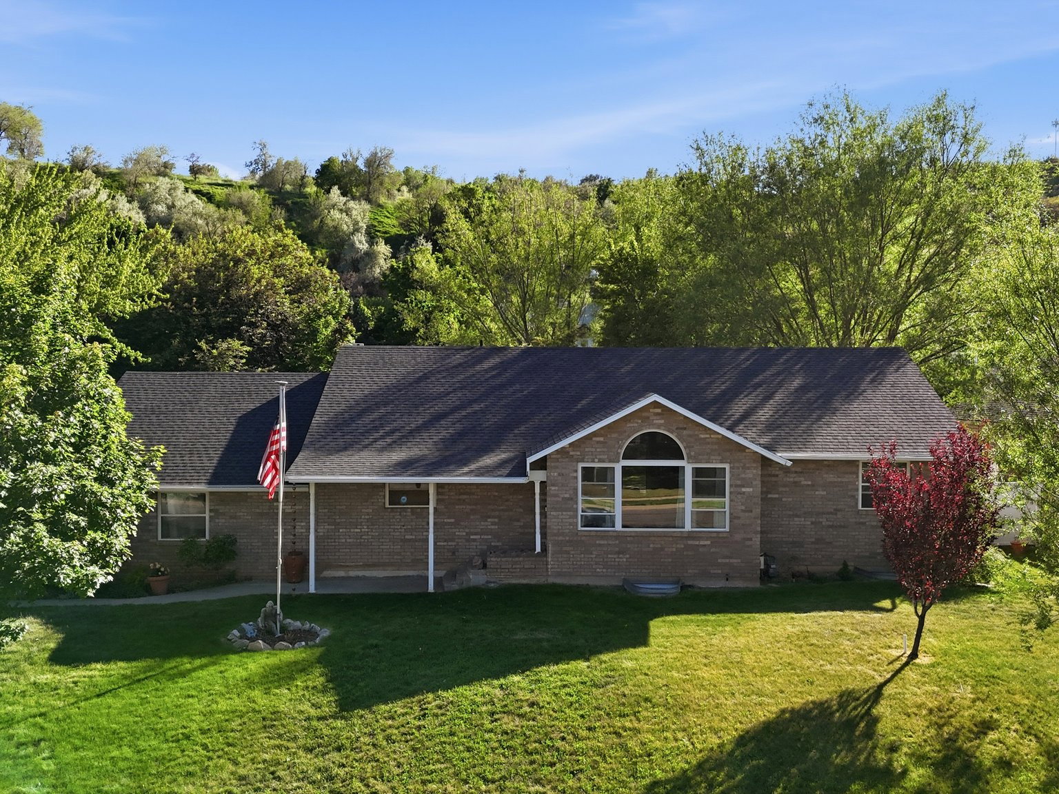 Brick facade residence featuring a covered front porch, arched window, dark shingle roof, and mature perimeter trees
